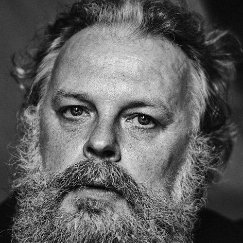 A man with a full beard and wavy hair in a black shirt